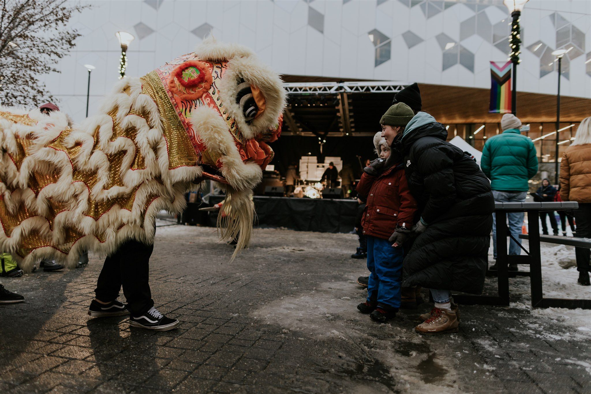 Traditional Chinese lion dance performer in white and gold costume interacting with bundled-up spectators at outdoor winter event, with stage and geometric wall pattern visible in background