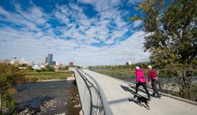 Two pedestrians and a dog crossing the Elbow River Traverse pedestrian bridge, showing downtown Calgary in the background