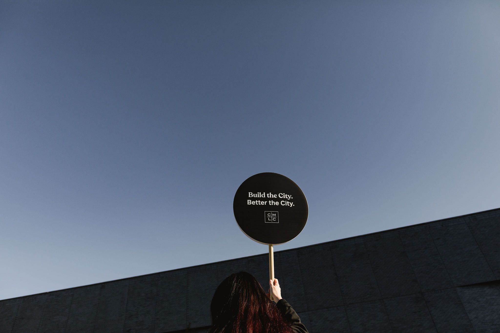 A person holding a sign that says "Built the City. Better the City." with the CMLC logo against a blue sky.