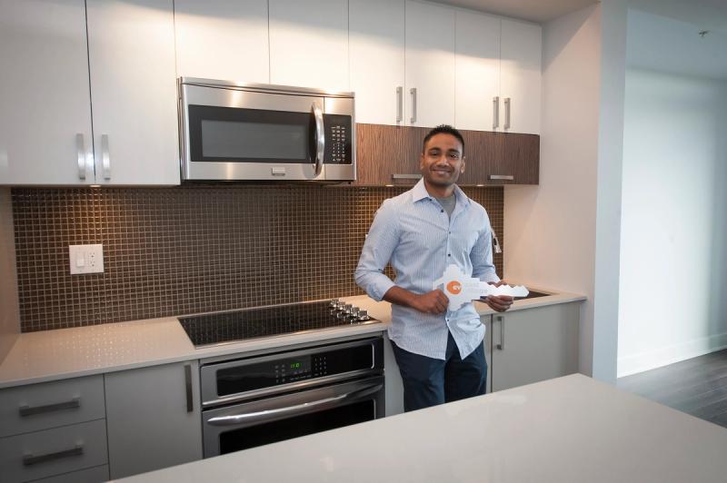 A photograph of 28 year-old pharmacist holding an oversized key with the East Village logo on it, smiling from his new kitchen in the FIRST residential tower