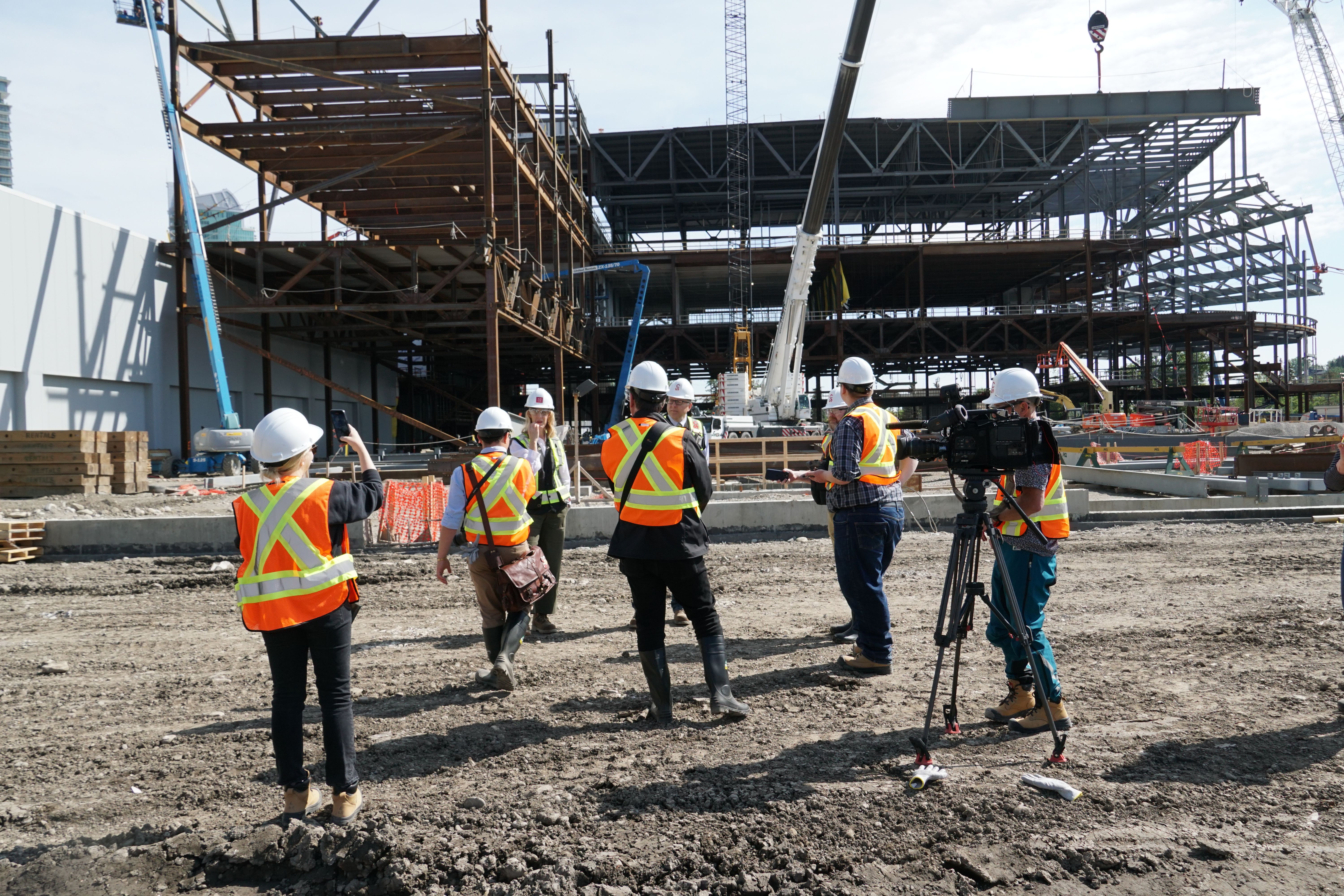 Construction site showing workers in safety gear (hard hats and high-visibility vests) documenting building progress with cameras and video equipment, with steel framework structure visible in background