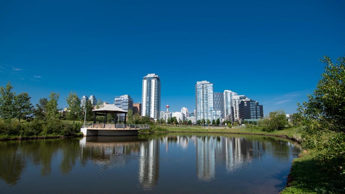 A photograph of the storm pond and band-stand style music pavilion in East Village. The photo shows trees and green space around the storm pond and the downtown Calgary skyline in the background.