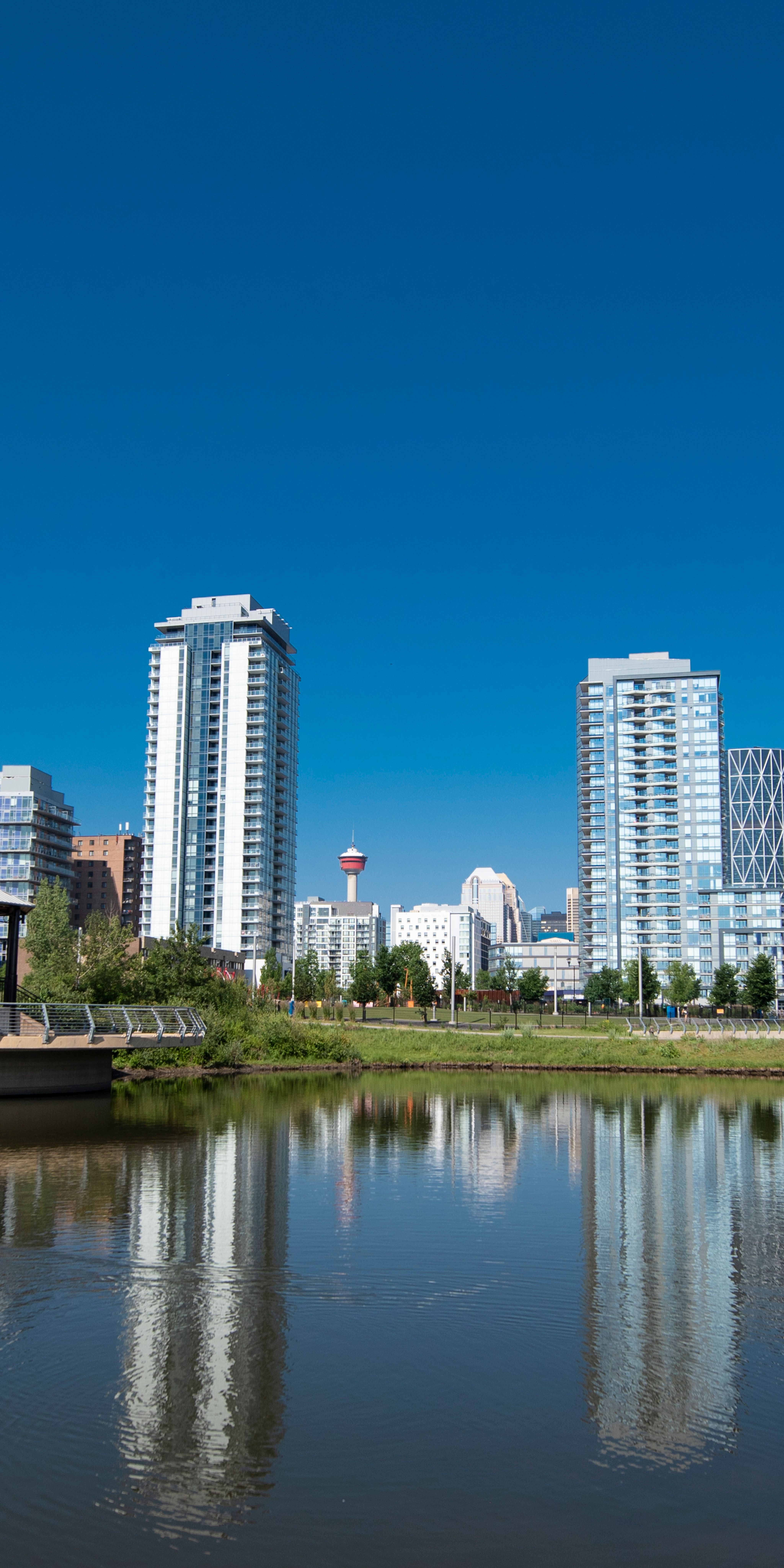 A photograph of the storm pond and band-stand style music pavilion in East Village. The photo shows trees and green space around the storm pond and the downtown Calgary skyline in the background.