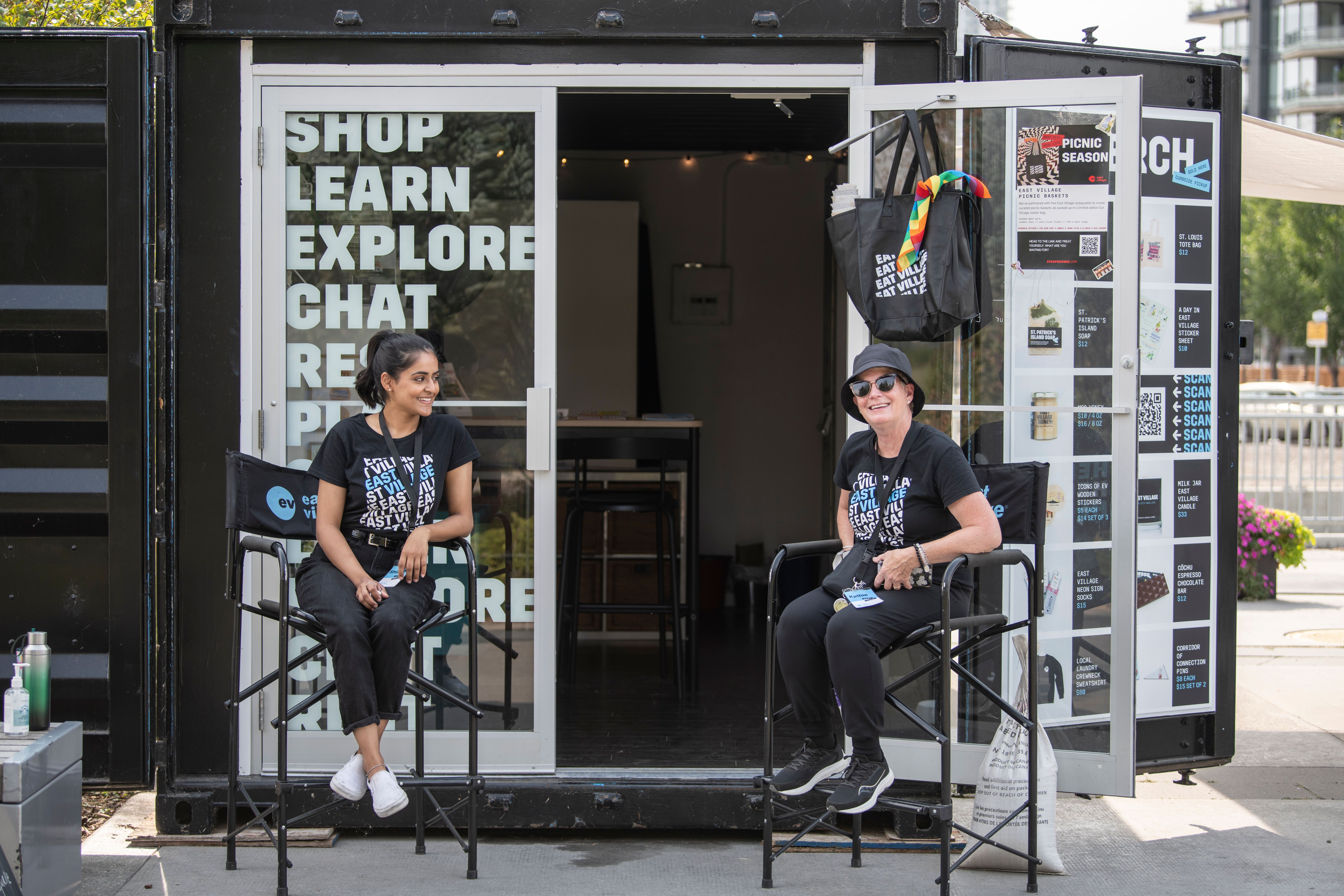 A photograph of two women sitting in front of the East Village HQ container.
