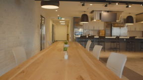 A photography of a long table with white chairs around it in the foreground and the commercial kitchen in the background