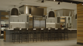 A photograph of a commercial kitchen, stainless steel appliances and an island with many black bar stools around it