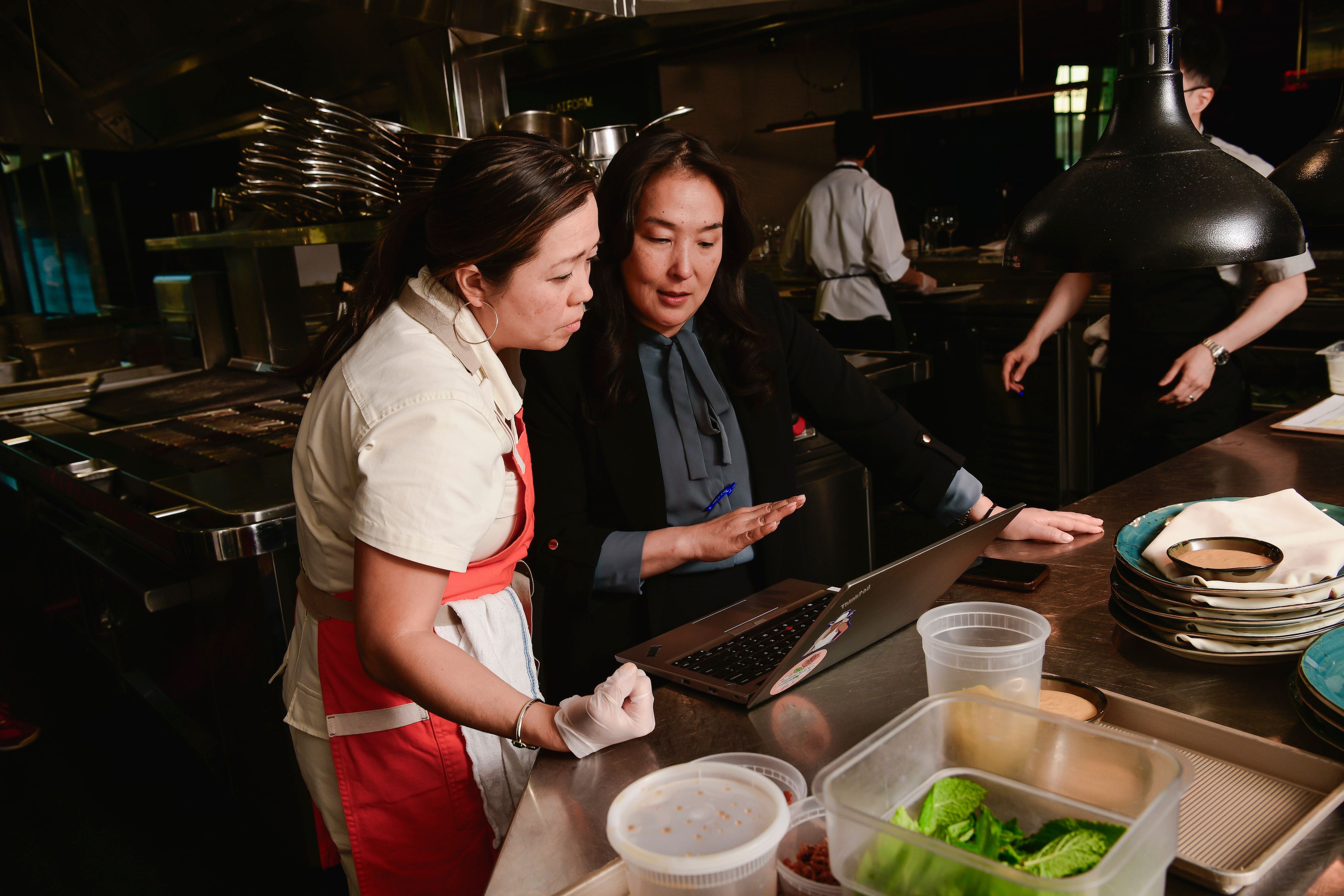 Two women stand around a computer in a restaurant kitchen