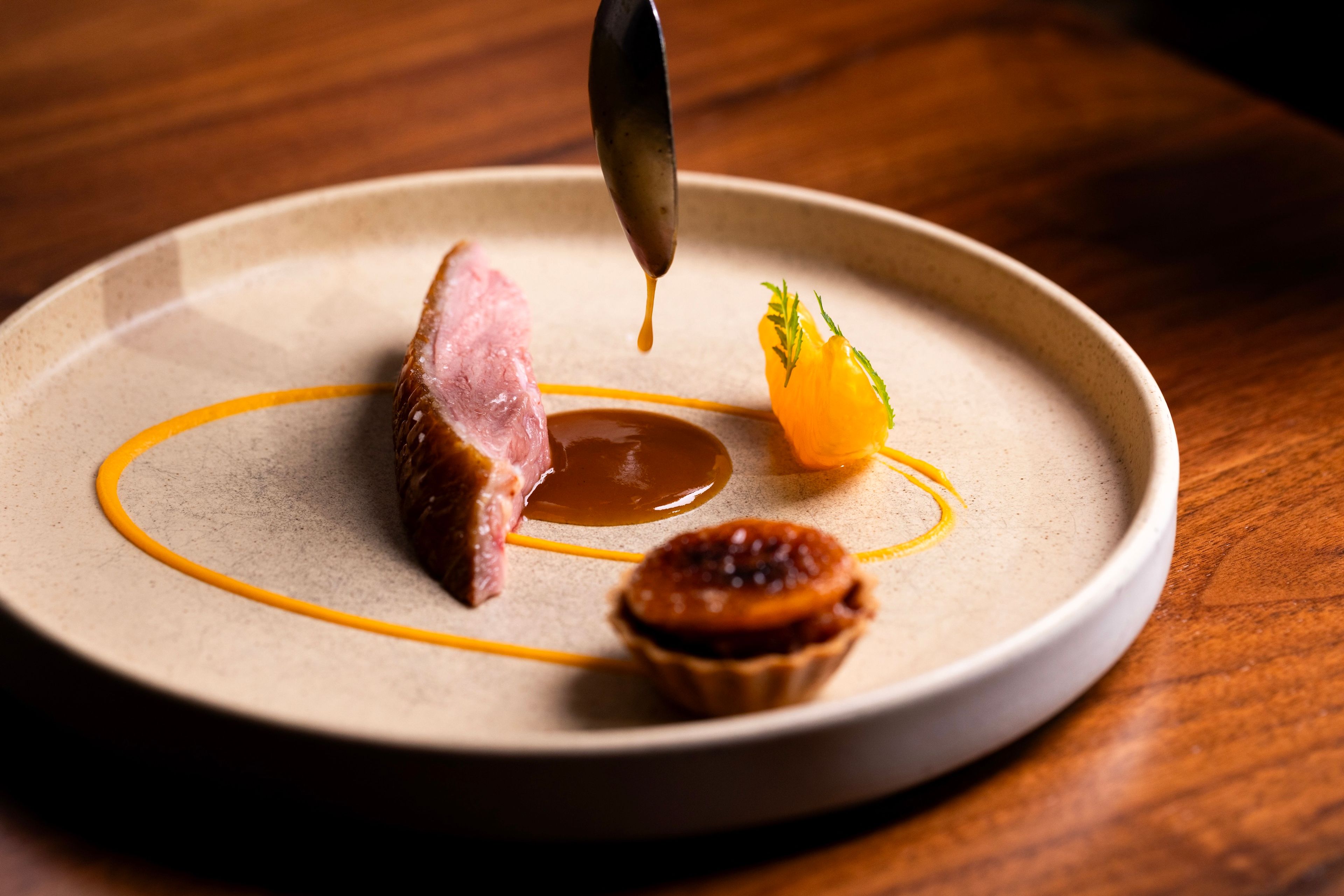 small piece of duck with a spoon dripping a brown sauce with other small components on a ceramic plate