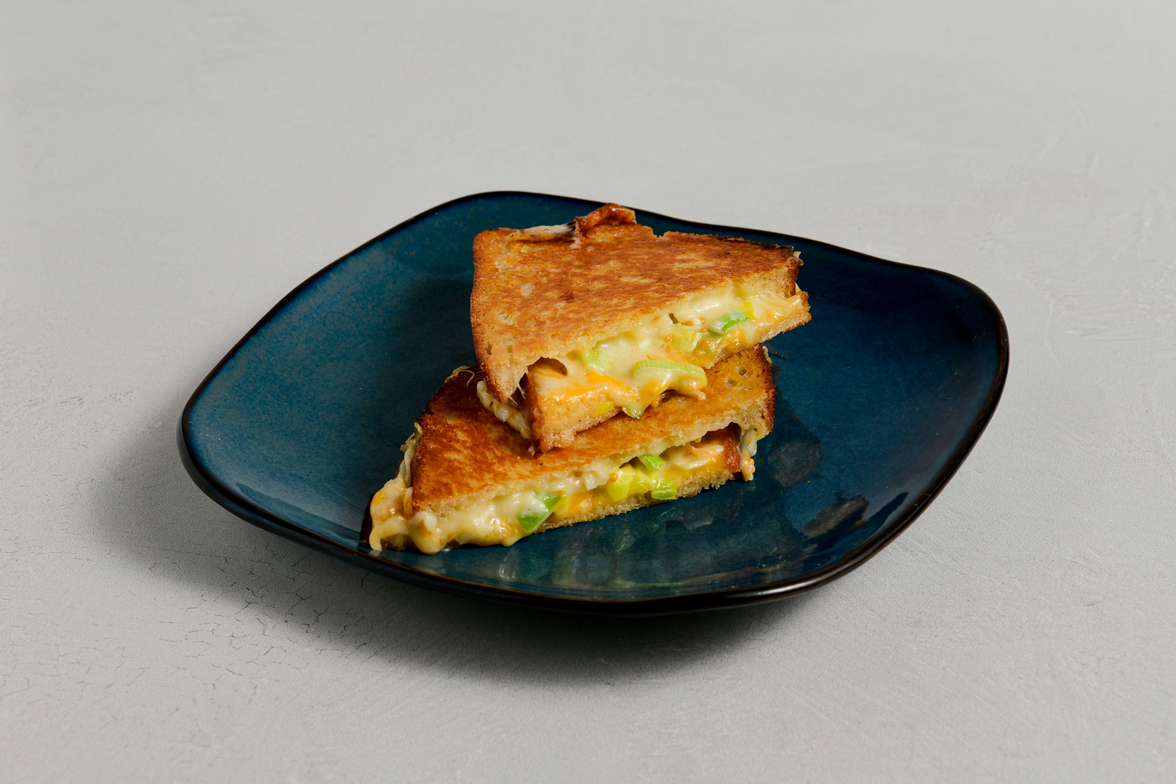 A grilled cheese with Bandaged Cheddar, Comté, Leek, and Pickled Mustard Seed on a blue plate
