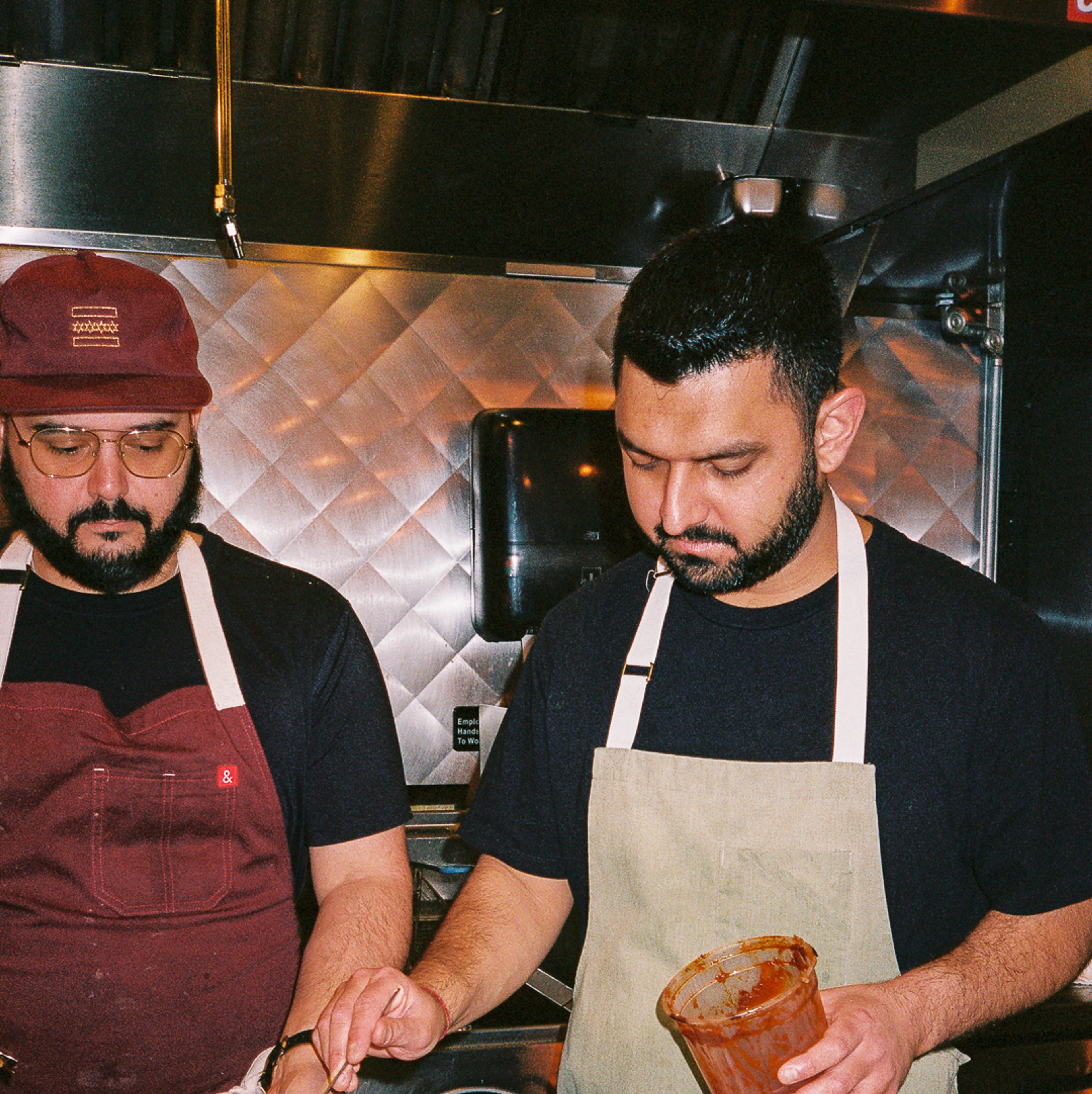 Zubair Mohajir holds a metal bowl in which Rishi Kumar working in a kitchen is placing a spoonful of red sauce
