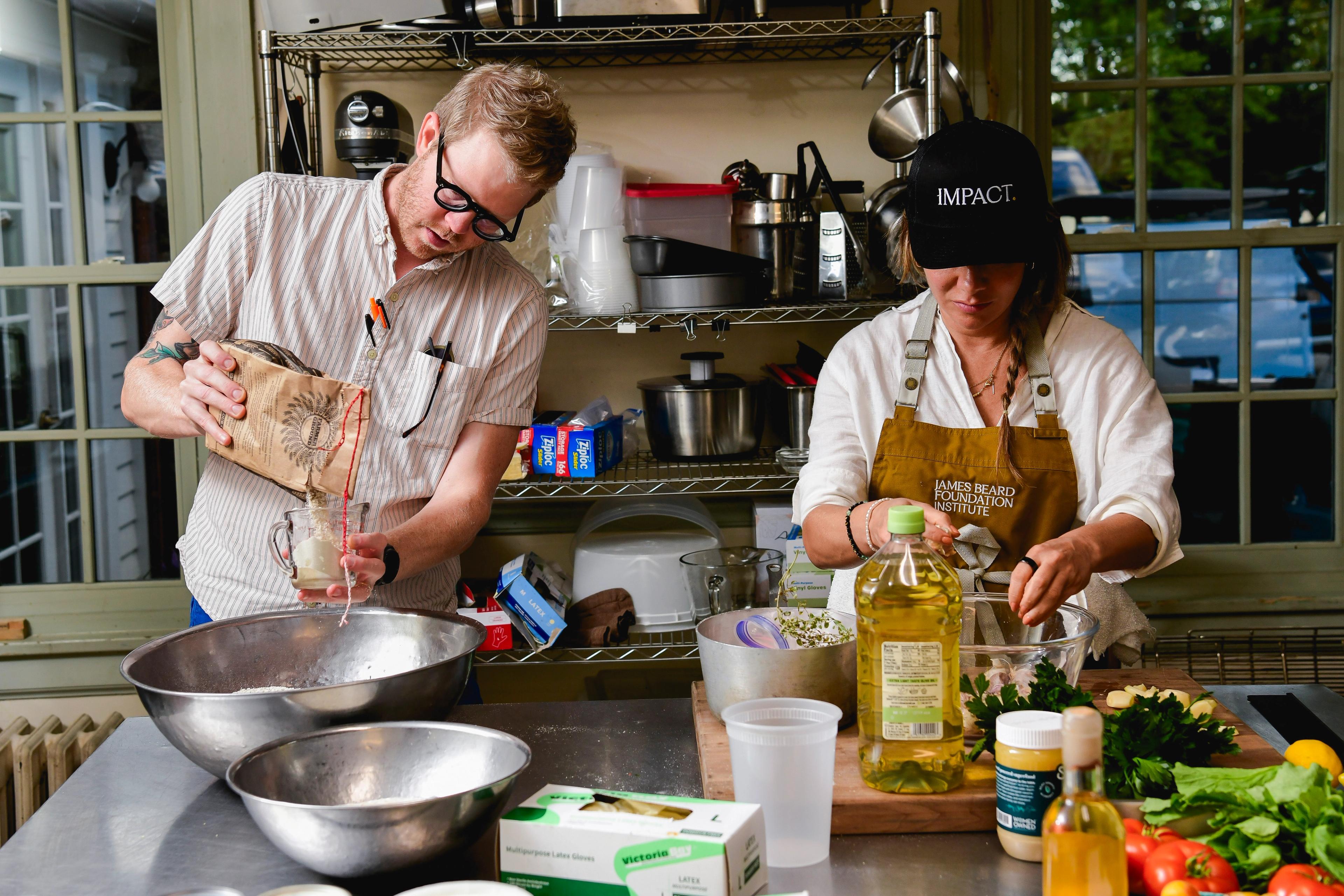 Two chefs work in a kitchen together. One is wearing an IMPACT. hat and a James Beard Foundation Institute apron.