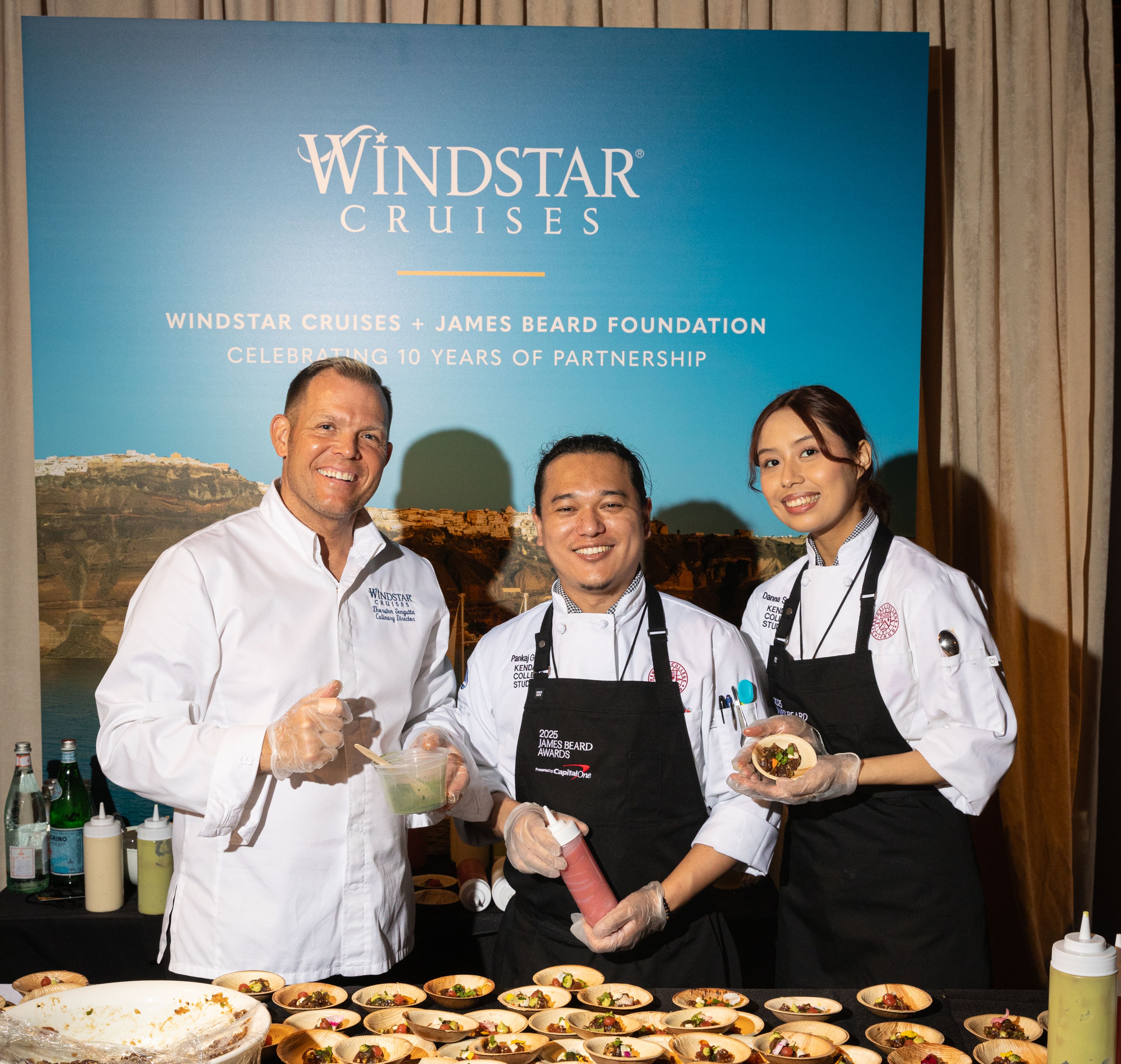 Three chefs pose in front of a backdrop featuring Windstar Cruises and the James Beard Foundation