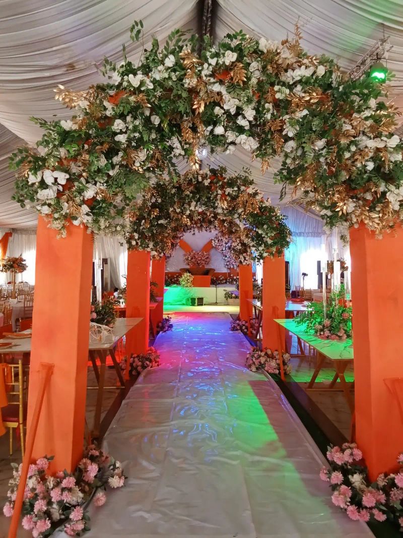 A flower covered arch over an orange draped aisle in a decorated hall