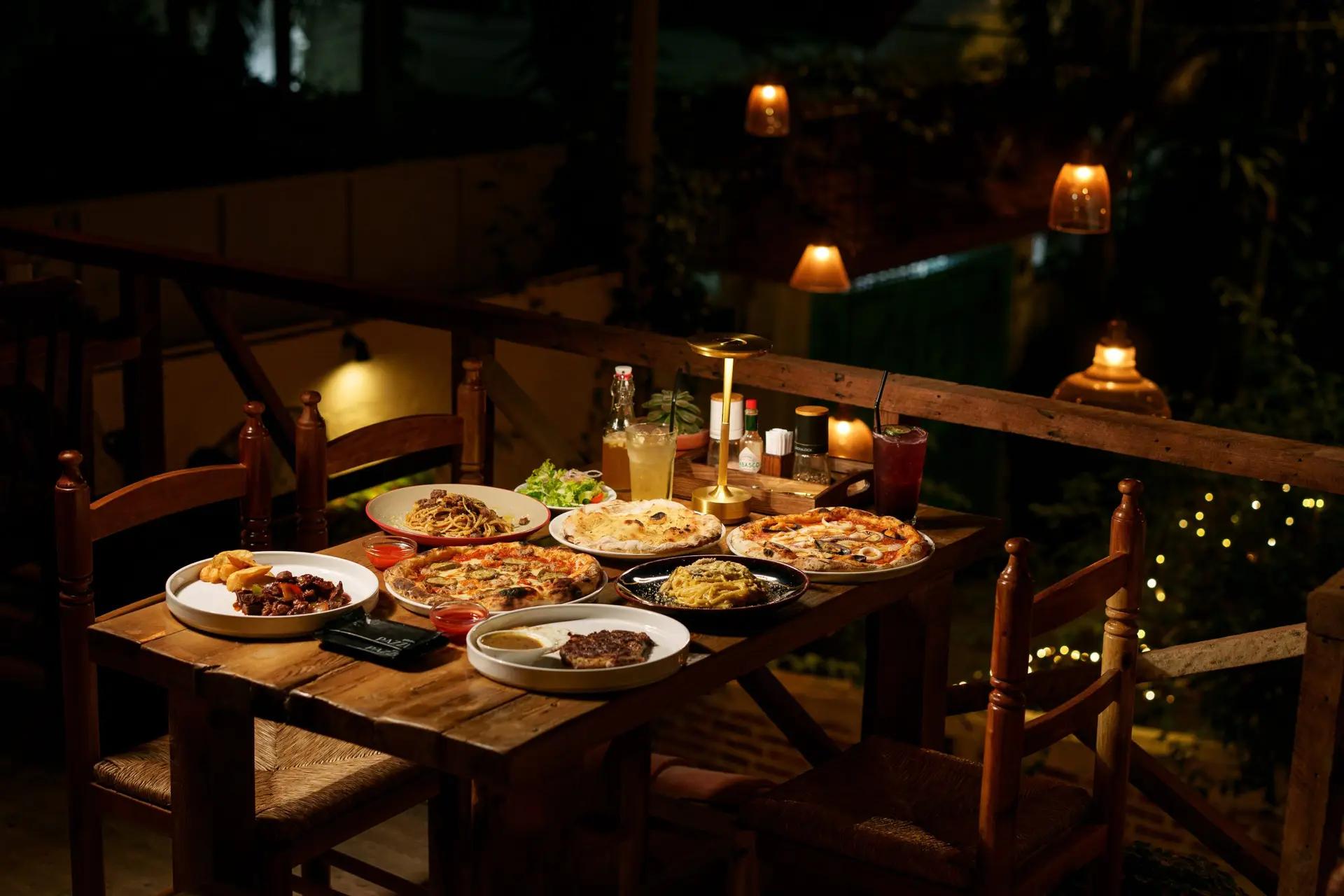 Bàn ăn tối lãng mạn tại nhà hàng pizza Ý Pazzi với nhiều chiếc pizza nóng hổi, pasta, món ăn kèm và đồ uống trong không gian ngoài trời ấm áp