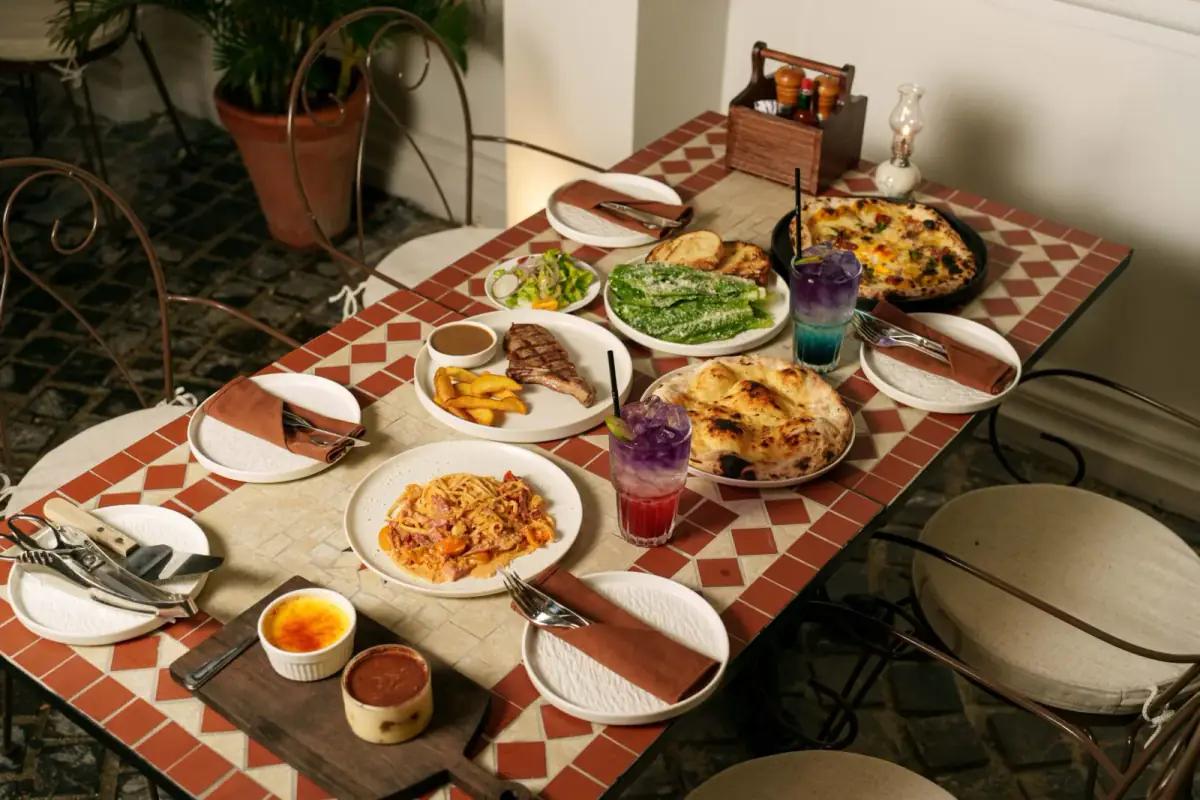 Bàn ăn tại nhà hàng pizza Ý Pazzi Quận 10 với pizza nướng, pasta, thịt nướng, salad tươi, đồ uống được sắp xếp trên khăn trải bàn kẻ đỏ trắng truyền thống