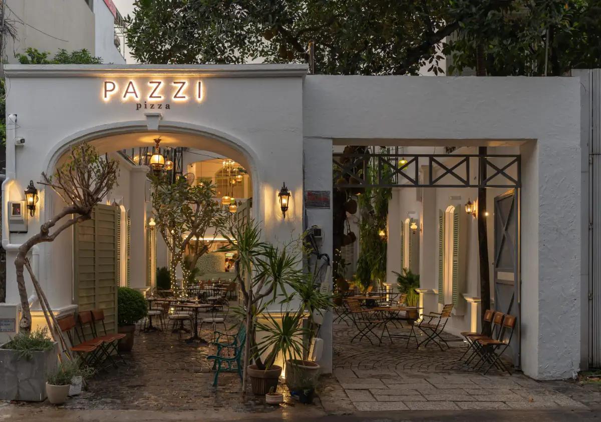 Mặt tiền nhà hàng pizza Pazzi Quận 10 với kiến trúc hiện đại, tường trắng, biển hiệu neon, cửa vòm, sân vườn xanh tươi và ánh sáng ấm áp buổi tối
