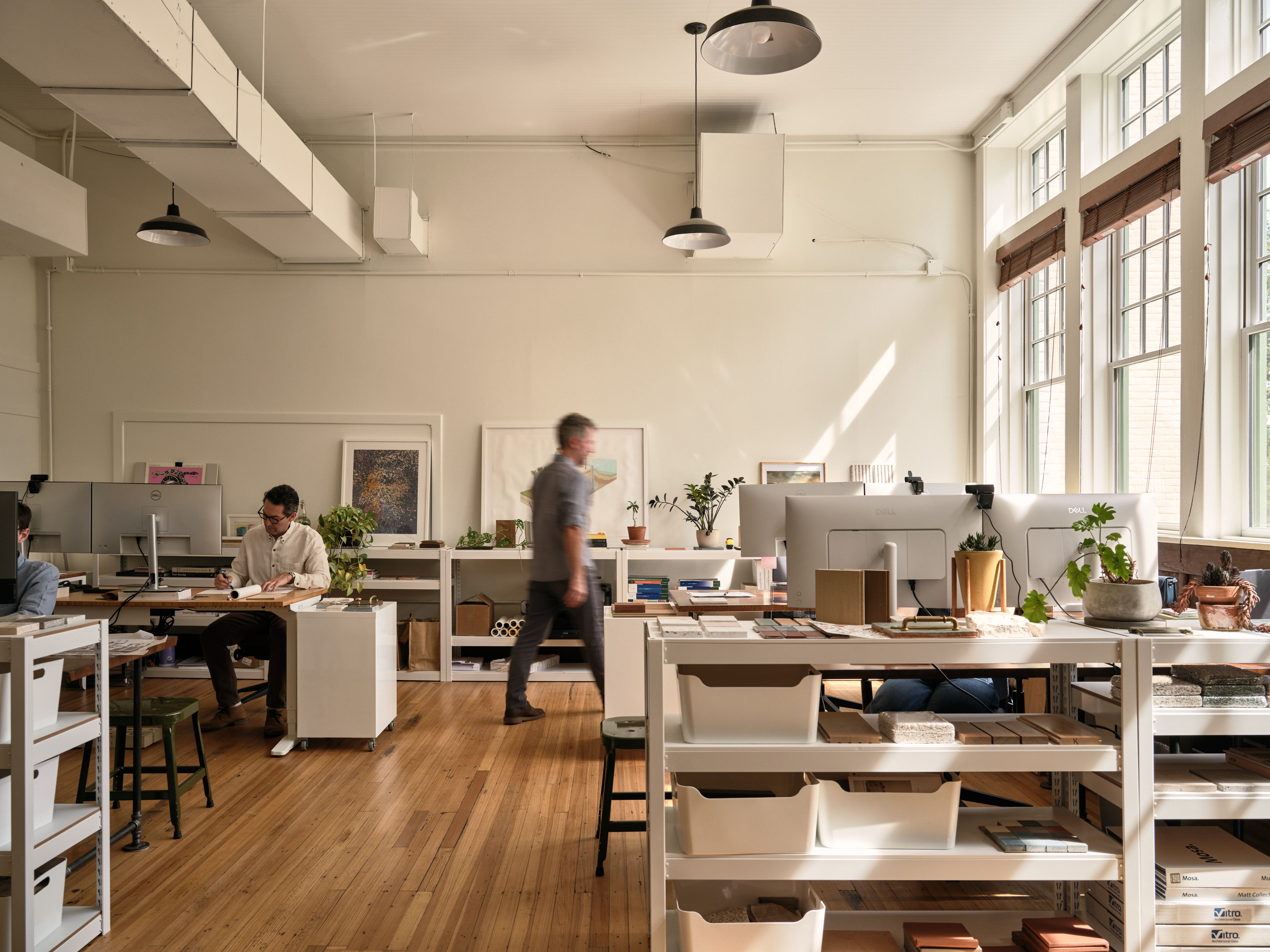 Nolan Bell's interior office space. People working and walking through a room with computers, desks and material samples. Large windows along the righthand wall.