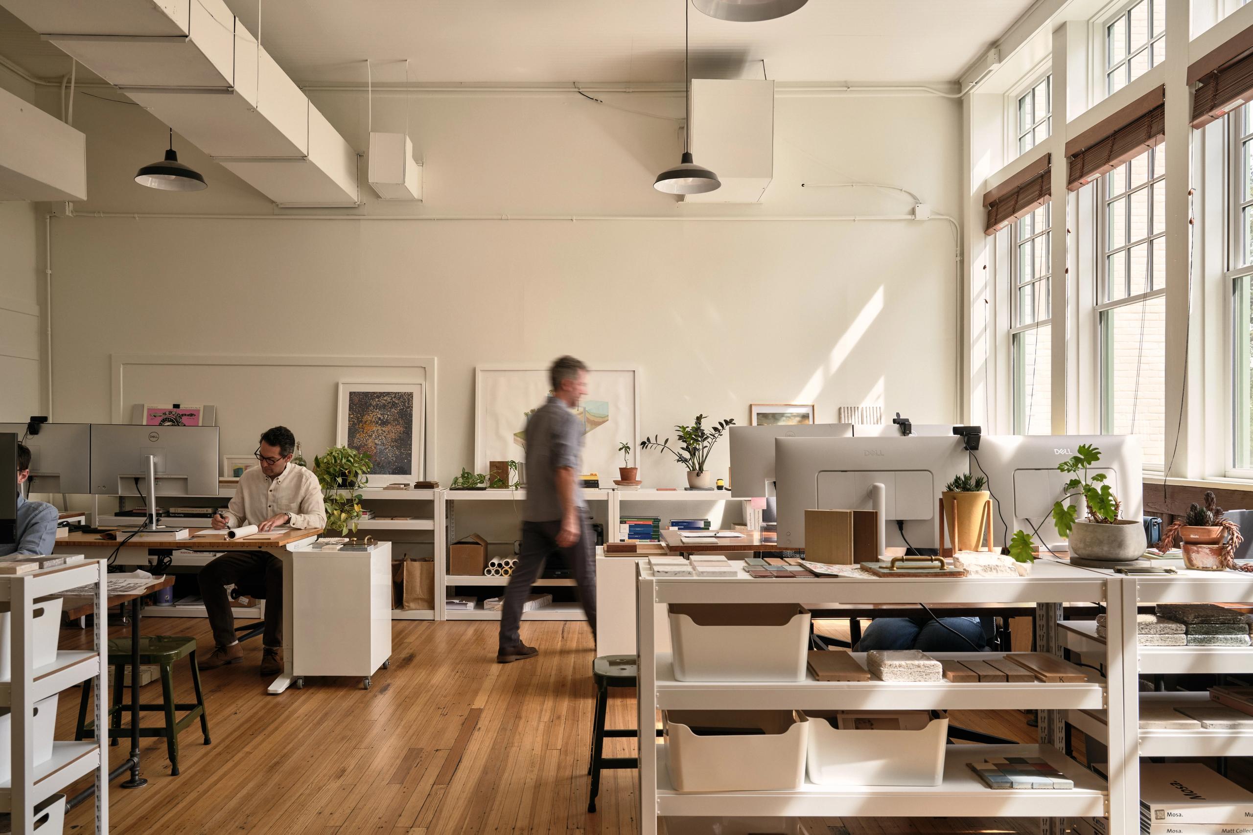 Nolan Bell's interior office space. People working and walking through a room with computers, desks and material samples. Large windows along the righthand wall.