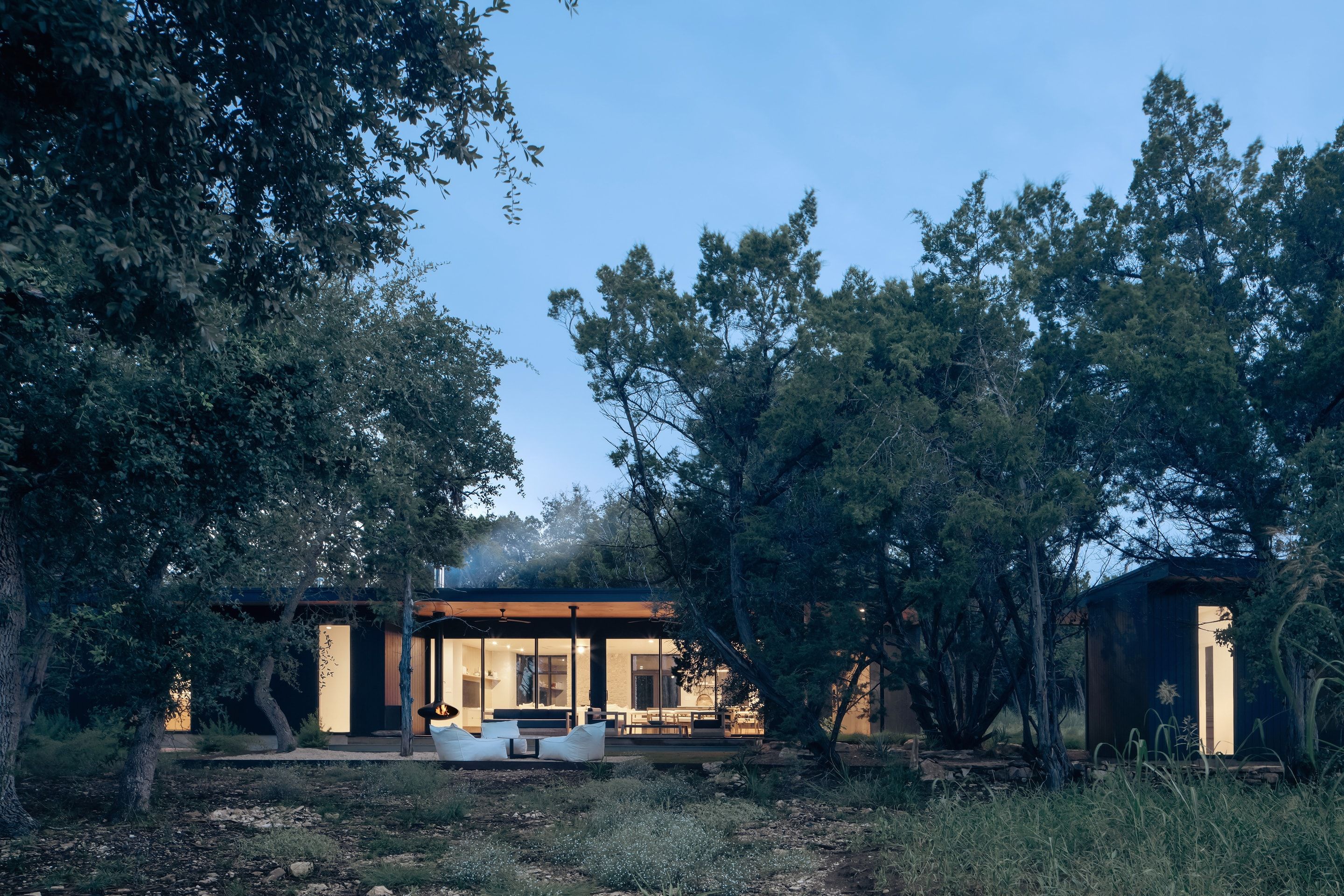 Low house at dusk, set among trees with glass walls open to a lit interior