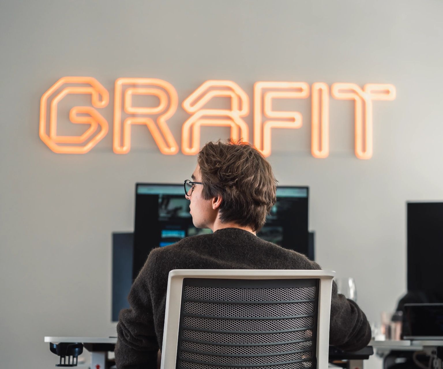 A team member working at a desk in the GRAFIT office, with the GRAFIT neon sign glowing on the wall behind them.