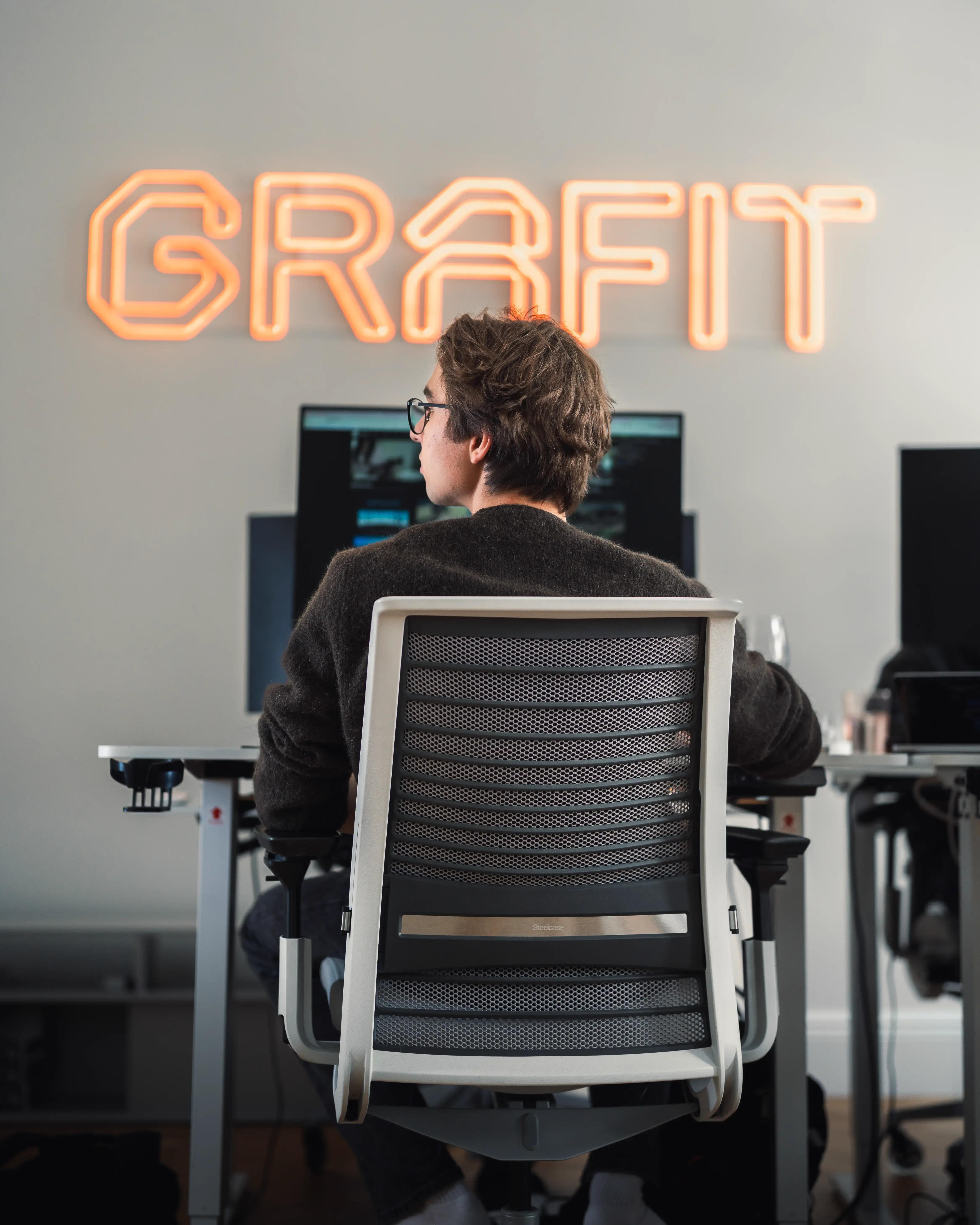 A team member working at a desk in the GRAFIT office, with the GRAFIT neon sign glowing on the wall behind them.