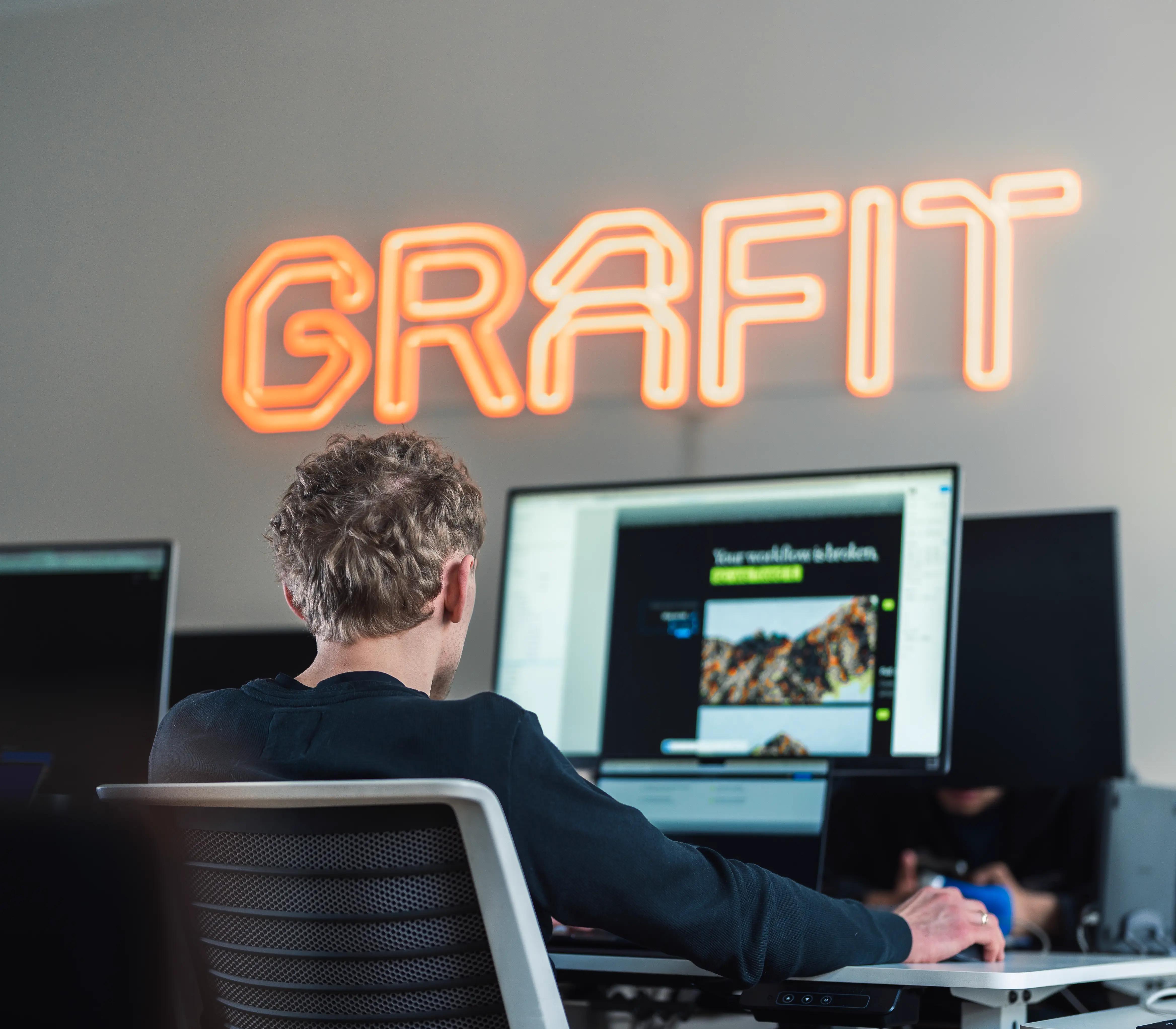 A team member working at a desk in the GRAFIT office, with the GRAFIT neon sign glowing on the wall behind them.