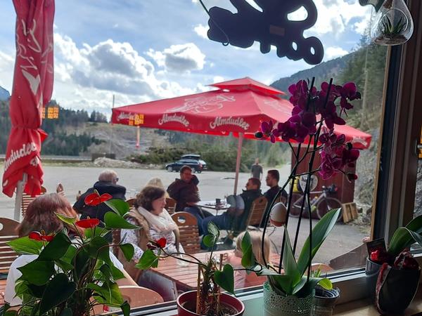 Blick durchs Fenster auf die sonnige Terrasse mit Gästen und Bergpanorama