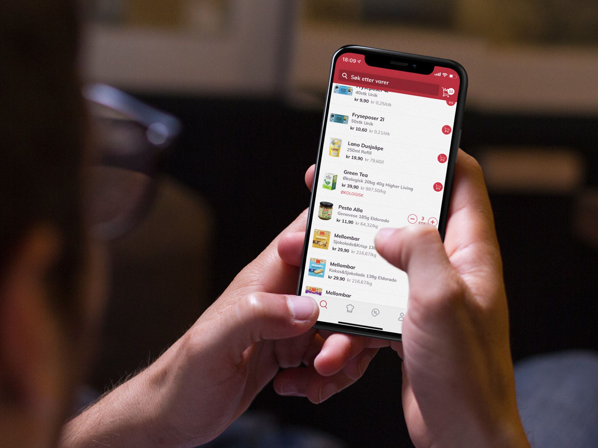 En person holder en smarttelefon som viser en app med ulike menyer og ikoner, sannsynligvis en mat- eller dagligvareapp.