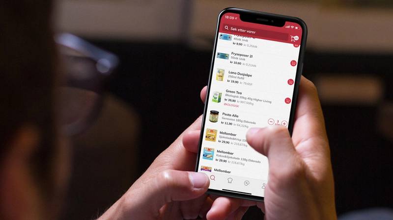 En person holder en smarttelefon som viser en app med ulike menyer og ikoner, sannsynligvis en mat- eller dagligvareapp.