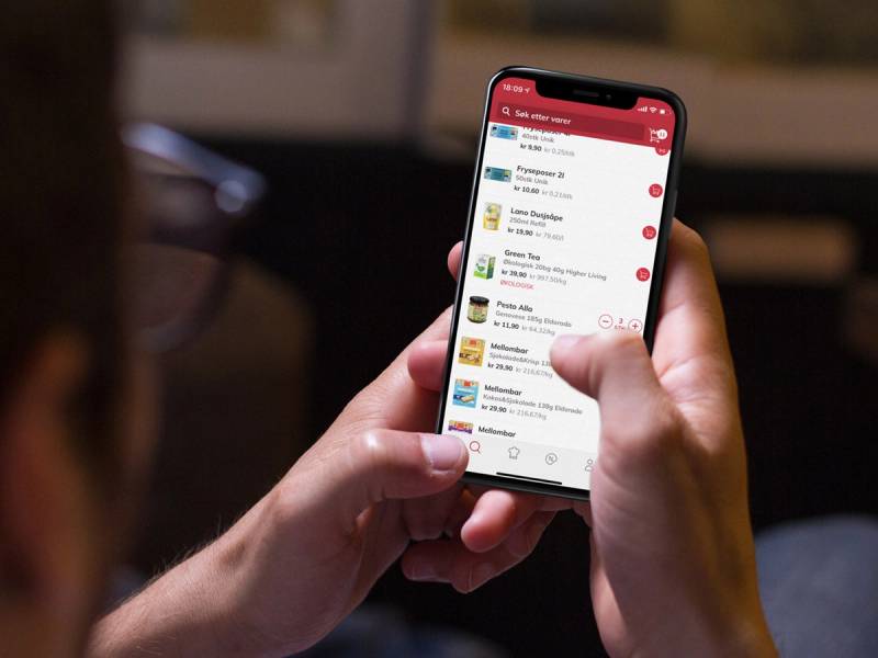 En person holder en smarttelefon som viser en app med ulike menyer og ikoner, sannsynligvis en mat- eller dagligvareapp.