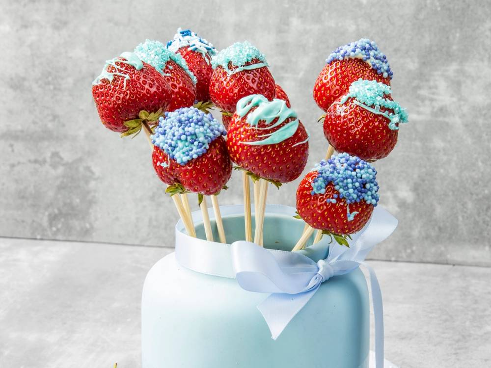 Fargerike jordbær på pinne med blå og røde kremdekorasjoner i en hvit vase, sannsynligvis til en babyshower.