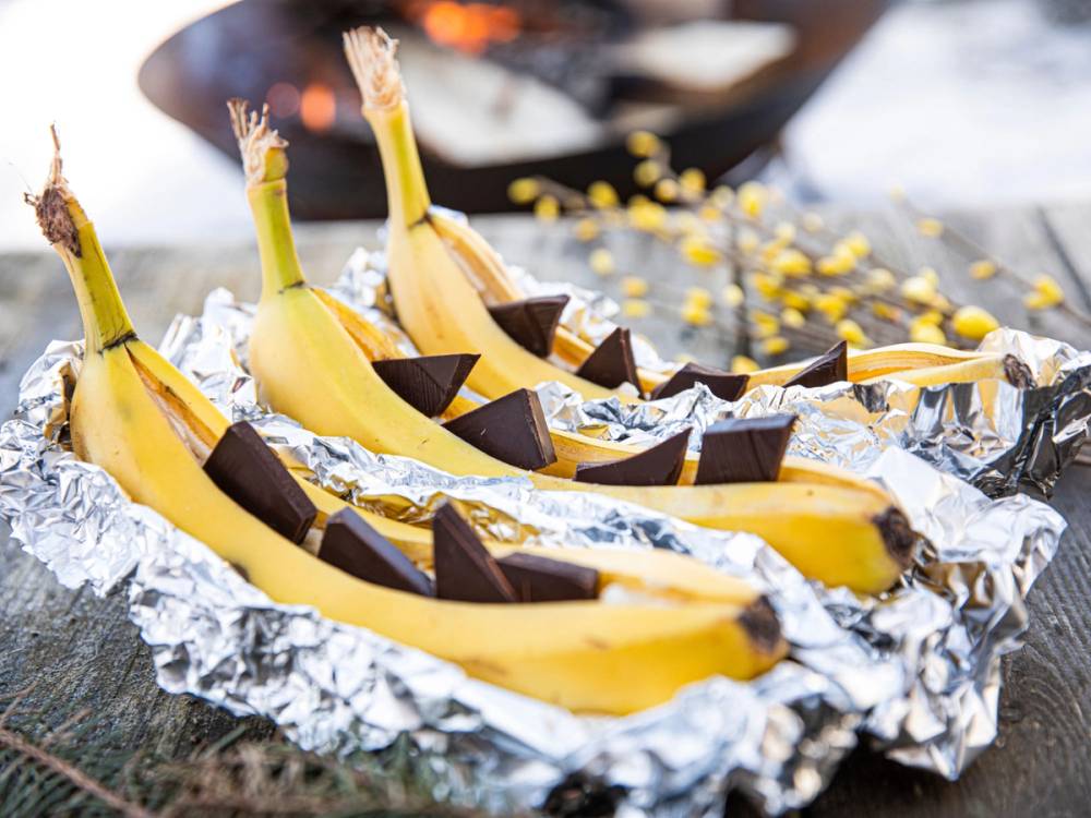 Bananer med sjokolade som er pakket inn i aluminiumsfolie og lagt på et bål for turmat, med en brennende ild i bakgrunnen.