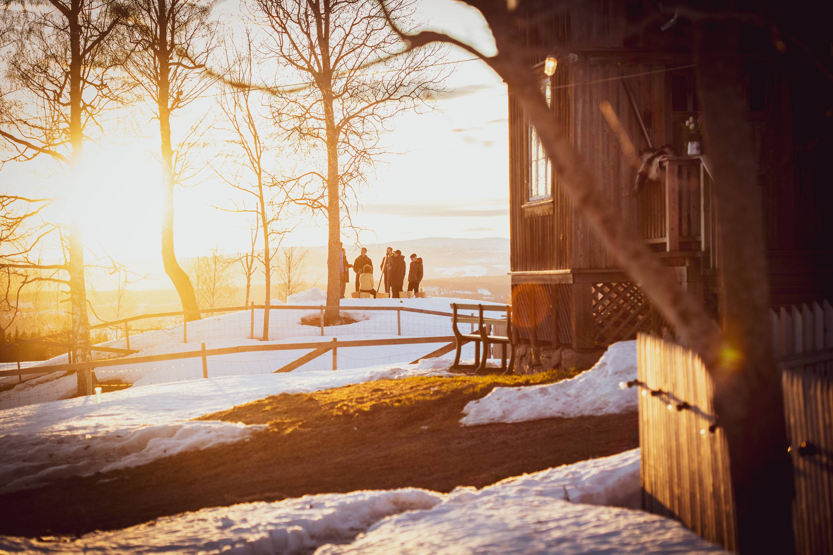 Solnedgang over en snødekt landskap med noen mennesker i bakgrunnen og en liten bygning til høyre.