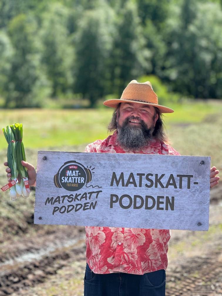 Person holder et skilt med teksten 'Matskatt-podden' og 'Matskatt-podden', med grønne planter i hånden, i et utendørs landskap med trær i bakgrunnen.