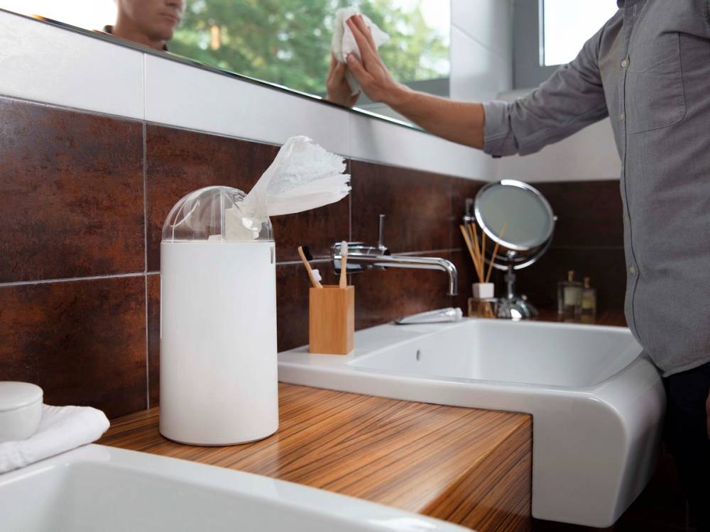En person tørker av et speil i et bad med en hvit håndkle, ved siden av en vask og en stor beholder med papirhåndklær.