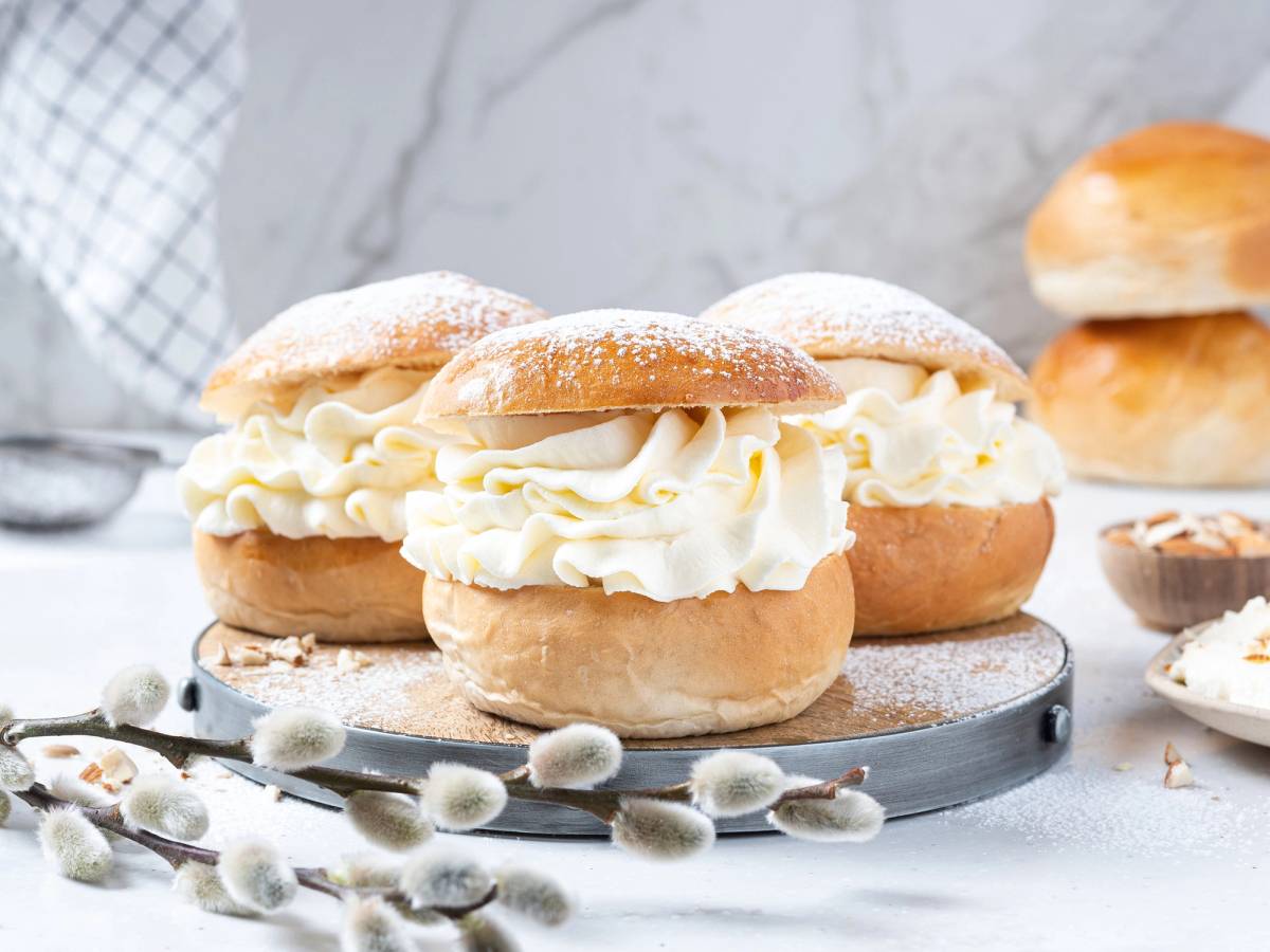 Fastelavnsboller fylt med krem og melis på et fat med blomster og bakverk i bakgrunnen.