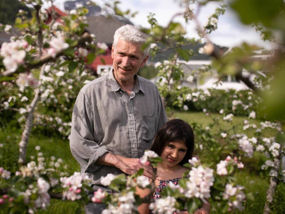 En person med grå skjorte står i en blomsterhage med en ung jente, omgitt av rosa blomster og trær, på en solrik dag.