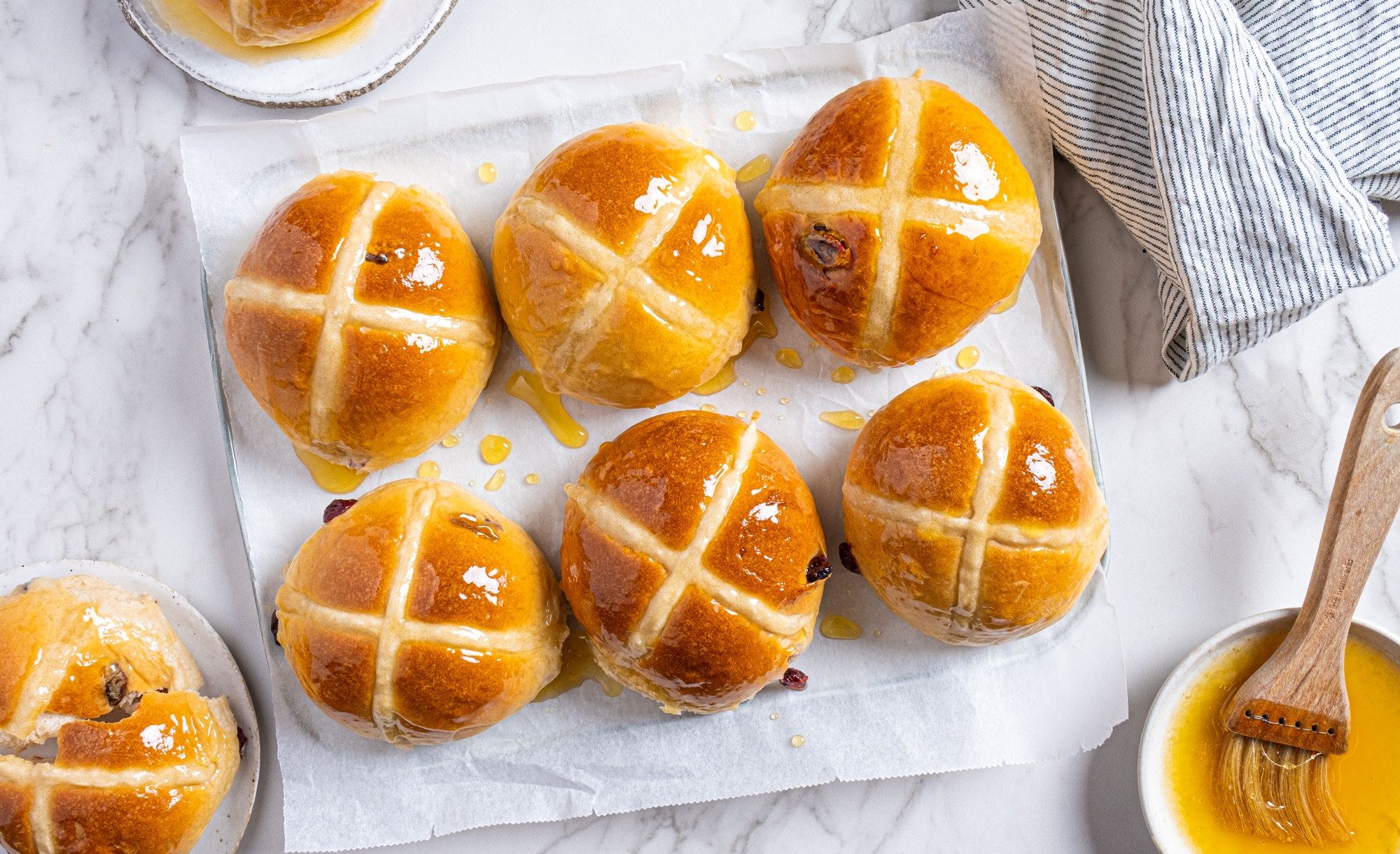 Syv varme hot cross buns med kryss av melisglasur på toppen, plassert på et hvitt brett med en liten skål med smør og en børste til å pensle med.