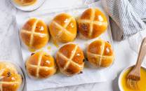 Syv varme hot cross buns med kryss av melisglasur på toppen, plassert på et hvitt brett med en liten skål med smør og en børste til å pensle med.