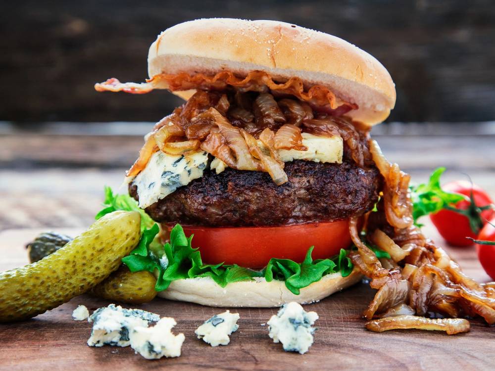 En hamburger med blåmuggost, kjøttkake, bacon, tomat, salat og sylteagurk på en treplate.