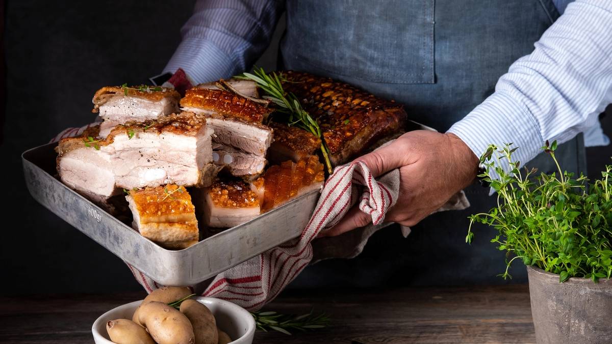 Person som holder en stor tallerken med ribbe, klar til servering, med poteter og grønne urter på bordet.