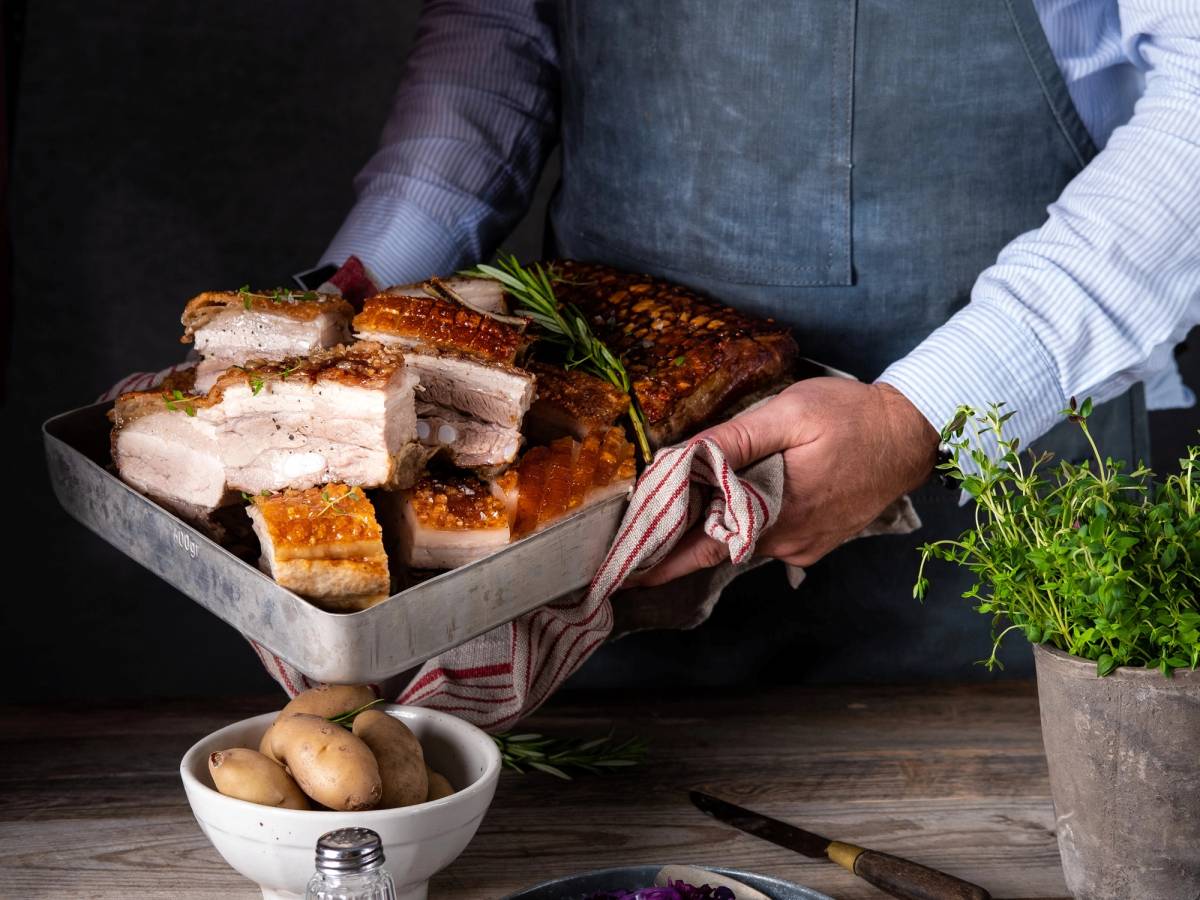 Person som holder en stor tallerken med ribbe, klar til servering, med poteter og grønne urter på bordet.