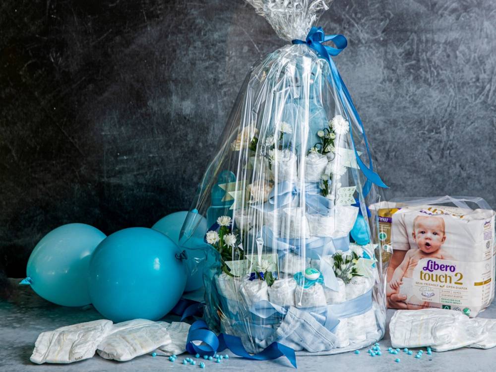 En bleiekake dekorert med hvite blomster og blå bånd, omgitt av blå ballonger og bleiepakkepakker, satt opp mot en mørk bakgrunn.