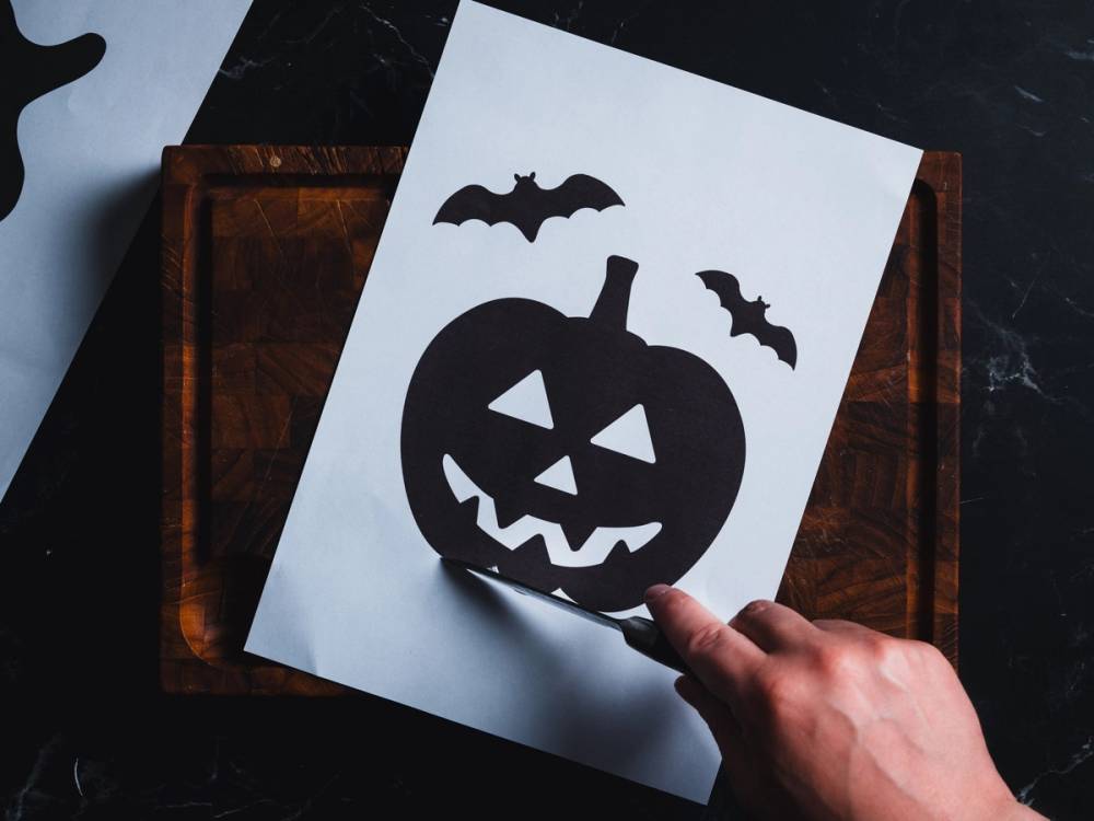 En person lager en halloween-utskjæring av en gresskar med ansikt, omgitt av to flaggermus, på et hvitt ark som ligger på en trebordflate.
