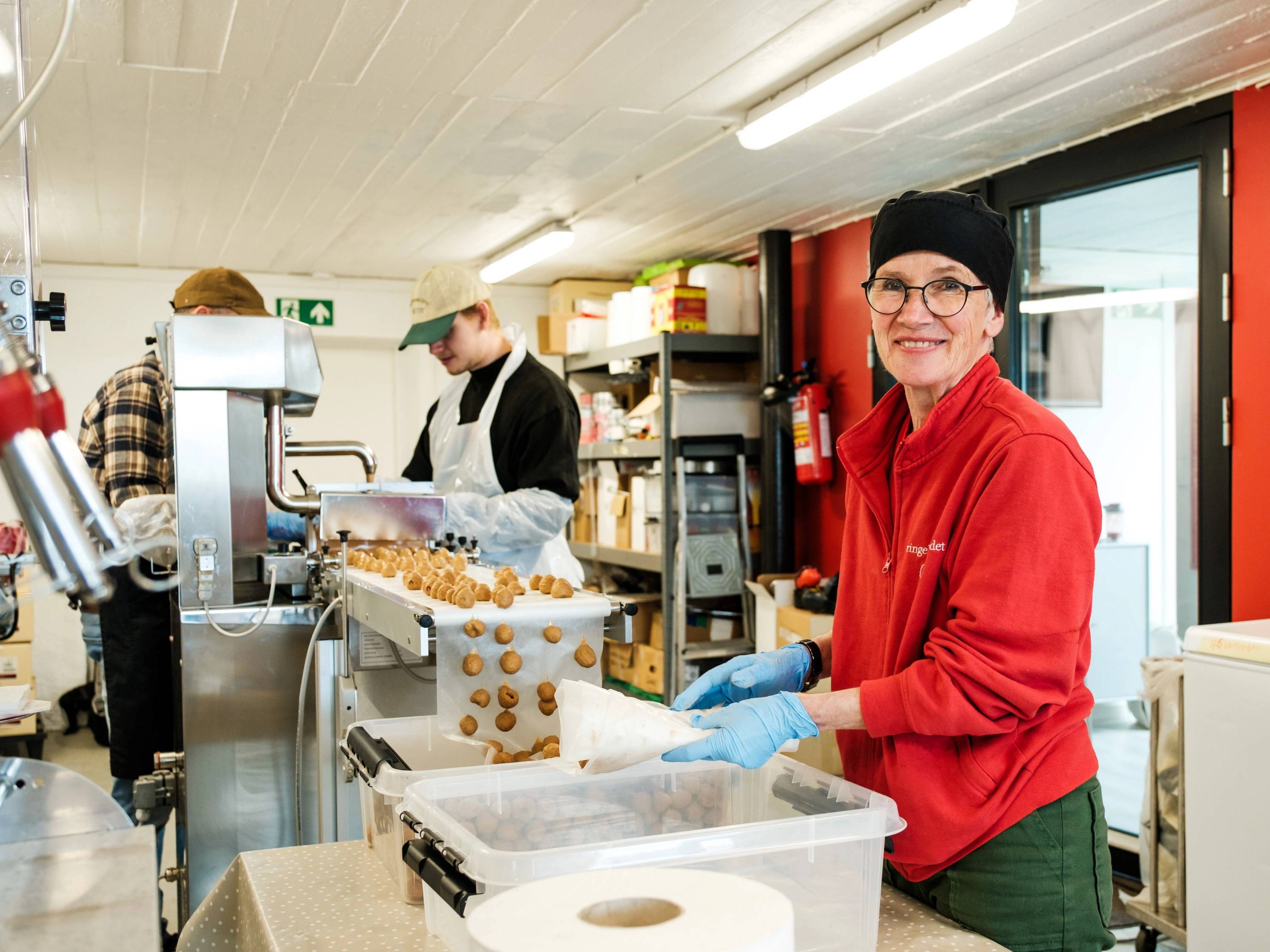 En person i rødt antrekk og blå hansker jobber med å pakke bringebær i en butikk eller på et marked.