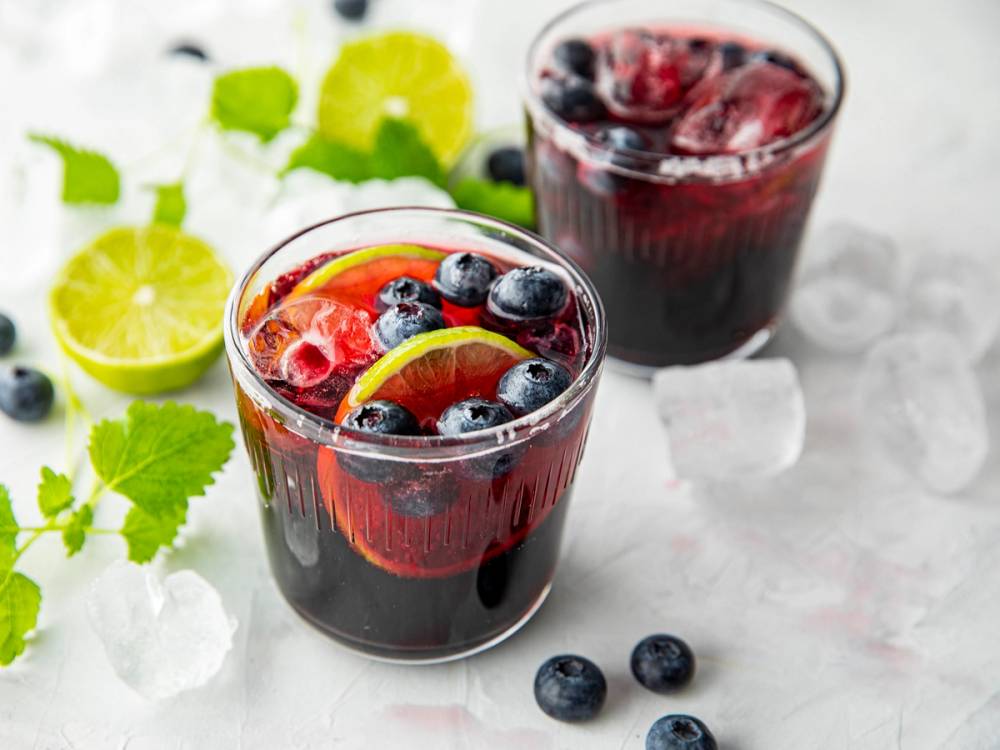 To glass med blåbærsaft med bær og sitronskiver, omgitt av sitronbåter og mynteblader, på en hvit overflate.