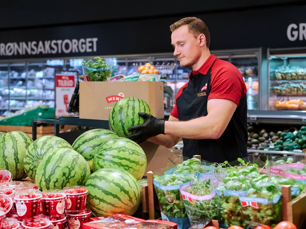 En person i en butikk med vannmeloner og andre fruktprodukter, iført rød og svart uniform, foran en kjøledisk med ulike varer.