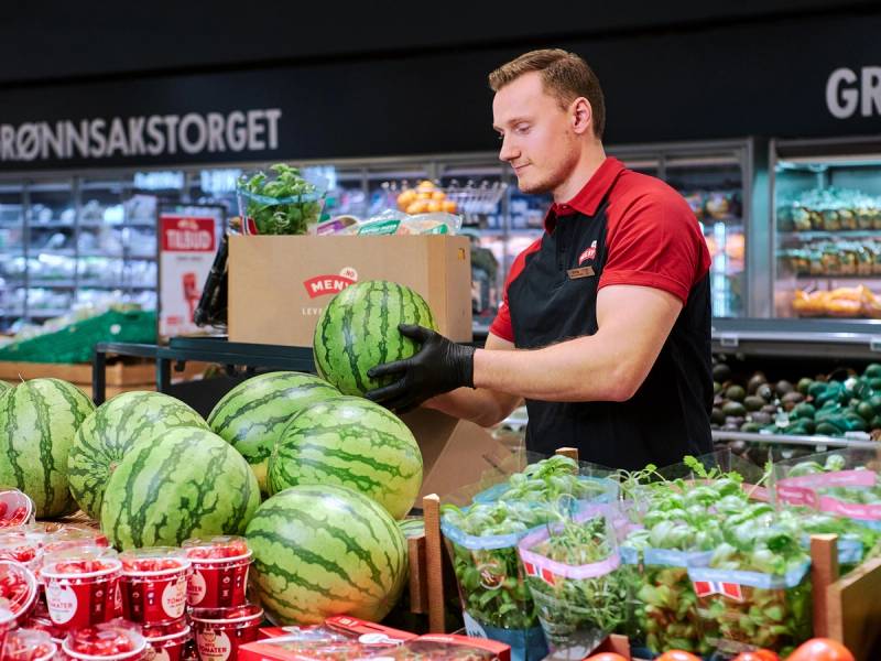 En person i en butikk med vannmeloner og andre fruktprodukter, iført rød og svart uniform, foran en kjøledisk med ulike varer.