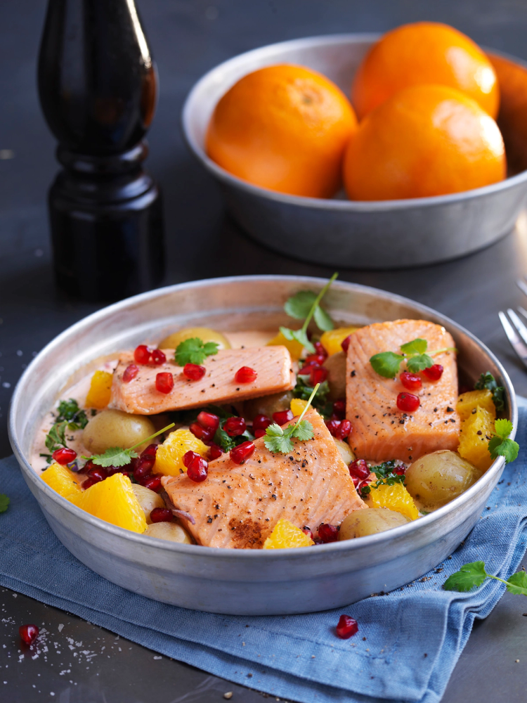 Laksefilet med creme fraiche og appelsin, pyntet med granateple og urter, servert i en rund hvit bolle med frukt i bakgrunnen.