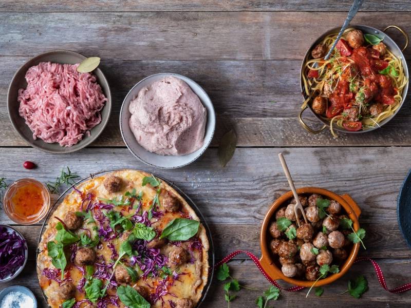 En pizza med grønnsaker og kjøttboller, omgitt av to typer medisterkaker og to typer saus, på et trebord.